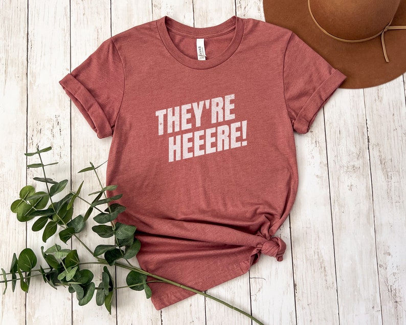 K&ouml;nnte beinhalten: Ein maronfarbenes T-Shirt mit dem Aufdruck "They're Heeeeere!" in Wei&szlig; auf der Vorderseite.