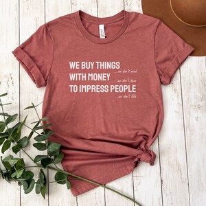 Może przedstawiać: Koszulka w kolorze bordowym z nadrukiem "WE BUY THINGS...we don't need WITH MONEY...we don't have TO IMPRESS PEOPLE...we don't like" w kolorze białym.