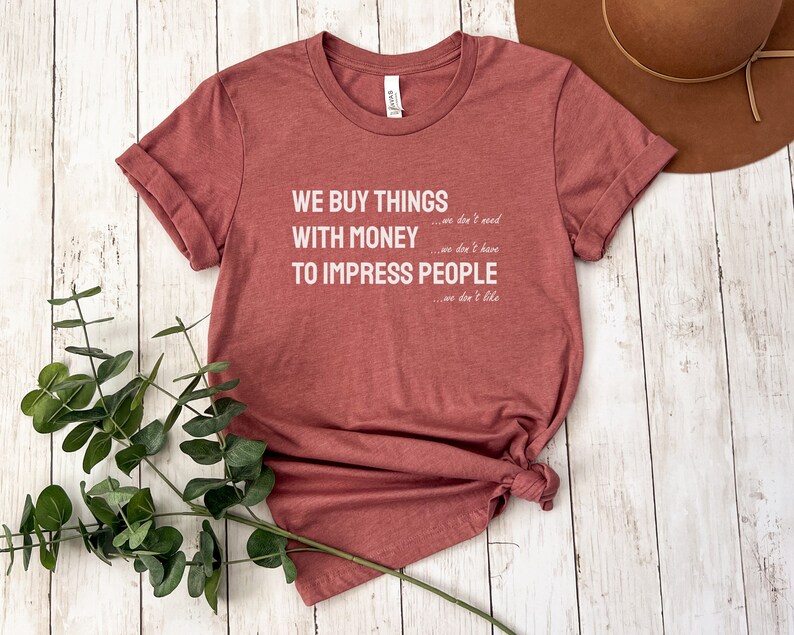Może przedstawiać: Koszulka w kolorze bordowym z nadrukiem "WE BUY THINGS...we don't need WITH MONEY...we don't have TO IMPRESS PEOPLE...we don't like" w kolorze białym.
