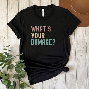 May include: Black t-shirt with the text "What's Your Damage?" in a retro font with pink, yellow, and blue colors.