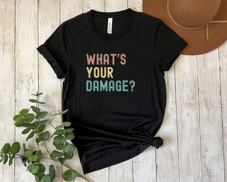 May include: Black t-shirt with the text "What's Your Damage?" in a retro font with pink, yellow, and blue colors.