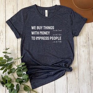 Może przedstawiać: Szary t-shirt z nadrukiem w kolorze białym, na kt&oacute;rym widnieje napis "WE BUY THINGS we don't need WITH MONEY we don't have TO IMPRESS PEOPLE we don't like".