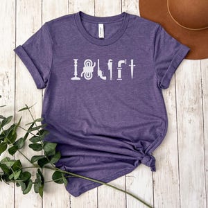 May include: Purple short-sleeved t-shirt with the text "I  LIFT" in white lettering. The text is made up of various tools, including a wrench, a hammer, a screwdriver, and a knife.