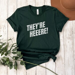 K&ouml;nnte beinhalten: Ein dunkelgr&uuml;nes T-Shirt mit dem Aufdruck "THEY'RE HEEEERE!" in Wei&szlig; auf der Vorderseite.