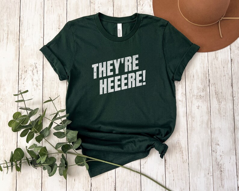 K&ouml;nnte beinhalten: Ein dunkelgr&uuml;nes T-Shirt mit dem Aufdruck "THEY'RE HEEEERE!" in Wei&szlig; auf der Vorderseite.
