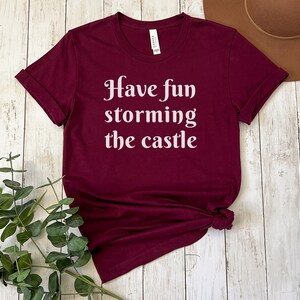 May include: A maroon t-shirt with the text "Have fun storming the castle" in white lettering.