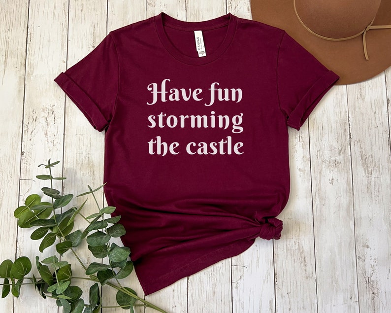 May include: A maroon t-shirt with the text "Have fun storming the castle" in white lettering.