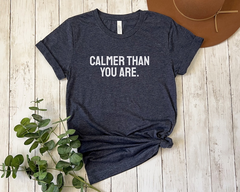 May include: A dark heather grey t-shirt with the text "Calmer than you are." printed in white.