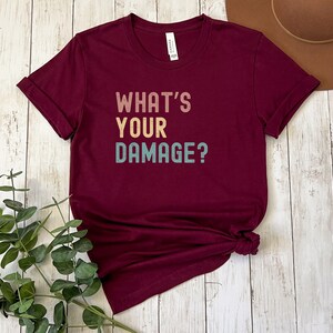 May include: A maroon t-shirt with the text "What's Your Damage?" in a vintage style font with colorful letters.