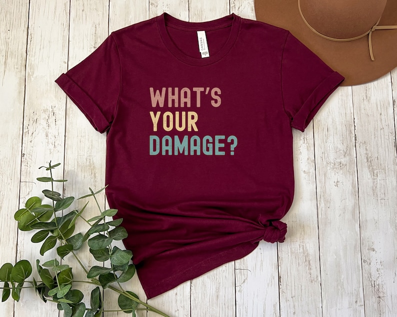 May include: A maroon t-shirt with the text "What's Your Damage?" in a vintage style font with colorful letters.