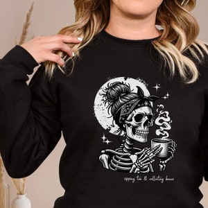 May include: Black and white illustration of a skeleton wearing a bandana, holding a cup of tea and a bone. The text reads "sipping tea & collecting bones".
