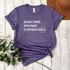 Może przedstawiać: Fioletowy T-shirt z nadrukiem "WE BUY THINGS we don't need WITH MONEY we don't have TO IMPRESS PEOPLE we don't like" w kolorze białym.