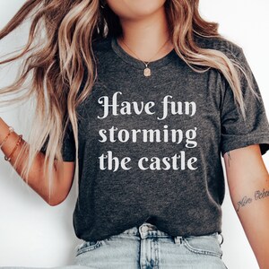 May include: A dark gray t-shirt with white text that reads "Have fun storming the castle".