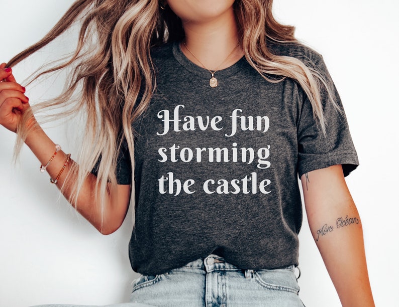 May include: A dark gray t-shirt with white text that reads "Have fun storming the castle".