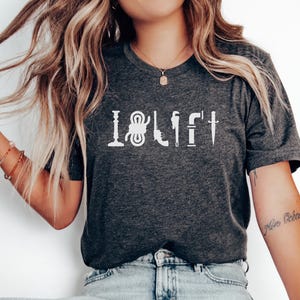 May include: A dark gray t-shirt with the text "I  LIFT" in white. The text is made up of various tools, including a wrench, a hammer, a rope, a pipe, and a knife.