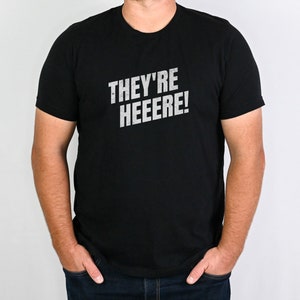 K&ouml;nnte beinhalten: Ein schwarzes T-Shirt mit dem Aufdruck "THEY'RE HEEEERE!" in Wei&szlig; auf der Vorderseite.