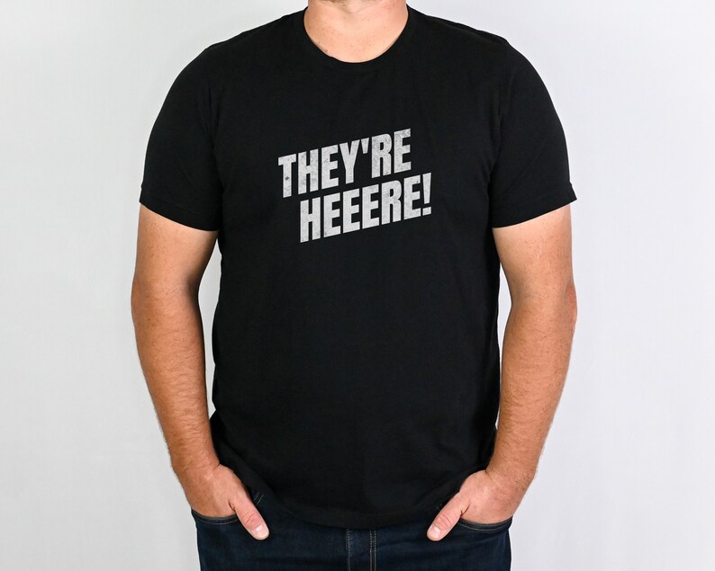 K&ouml;nnte beinhalten: Ein schwarzes T-Shirt mit dem Aufdruck "THEY'RE HEEEERE!" in Wei&szlig; auf der Vorderseite.
