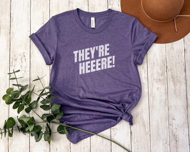 K&ouml;nnte beinhalten: Lilafarbenes T-Shirt mit kurzen &Auml;rmeln und dem Text "They're Heeeere!" in wei&szlig;en Buchstaben.