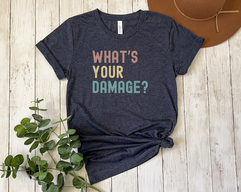 May include: A dark heather grey t-shirt with the text "WHAT'S YOUR DAMAGE?" in a retro font with colorful accents.