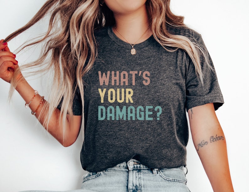 May include: A dark grey t-shirt with the text "WHAT'S YOUR DAMAGE?" printed in colorful letters. The text is in a retro style font.