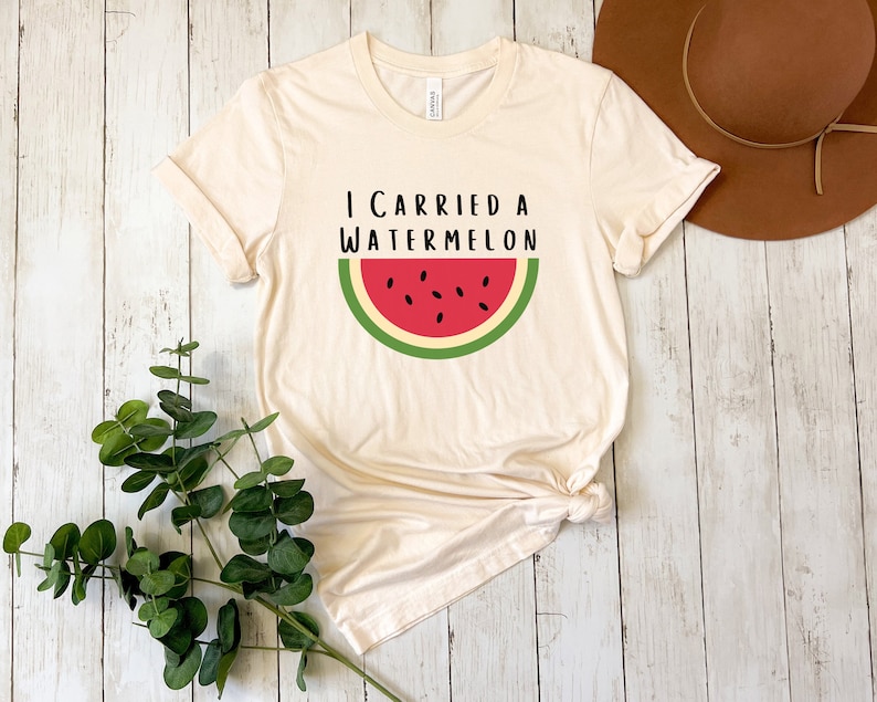 Może przedstawiać: Kremowa koszulka z nadrukiem "I carried a watermelon" i grafiką czerwonego plastra arbuza z zieloną sk&oacute;rką i czarnymi pestkami.