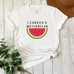 Może przedstawiać: Biała koszulka z napisem "I carried a watermelon" i grafiką czerwonego plastra arbuza z zieloną sk&oacute;rką i czarnymi pestkami.