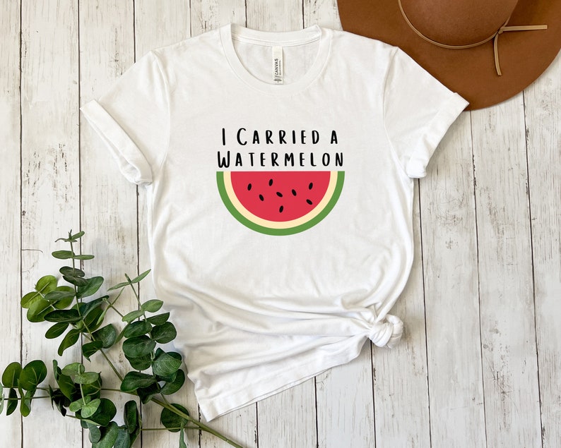 Może przedstawiać: Biała koszulka z napisem "I carried a watermelon" i grafiką czerwonego plastra arbuza z zieloną sk&oacute;rką i czarnymi pestkami.