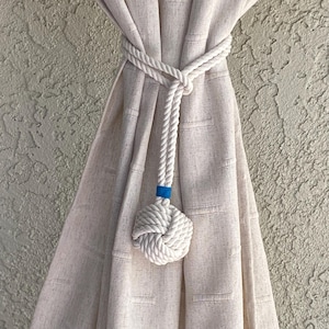 May include: White rope curtain tieback with a knotted ball at the end. The rope is tied around a beige curtain and the knot is accented with a small blue band.
