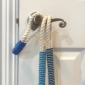 May include: A white rope door handle wrap with blue striped accents. The rope is wrapped around the door handle and is hanging down. The door handle is silver and the door is white.