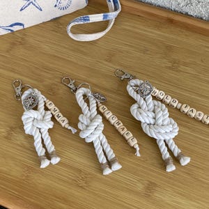 May include: Three white rope keychains featuring a nautical knot design, a silver-toned compass charm, and wooden beads. Two keychains are personalized with the names EMMA and MICHAEL, while the third features the text HOLD FAST.