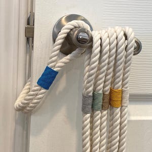 May include: A white rope door handle with a blue accent, looped around a silver door handle. Several strands of white rope hang down, each with a different colored accent: gray, light green, tan, yellow, and white. The door is white.