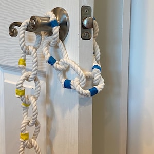 May include: A decorative rope chain with white rope and blue and yellow accents hangs from a door handle. The nautical-themed chain is made of thick, knotted rope, adding a touch of coastal charm to any space.