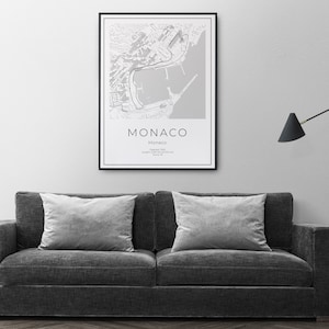 K&ouml;nnte beinhalten: Eine gerahmte Schwarz-Wei&szlig;-Karte von Monaco h&auml;ngt an einer grauen Wand &uuml;ber einem dunkelgrauen Sofa mit zwei grauen Kissen. Die Karte zeigt das Wort "Monaco" in einer klaren Schriftart.