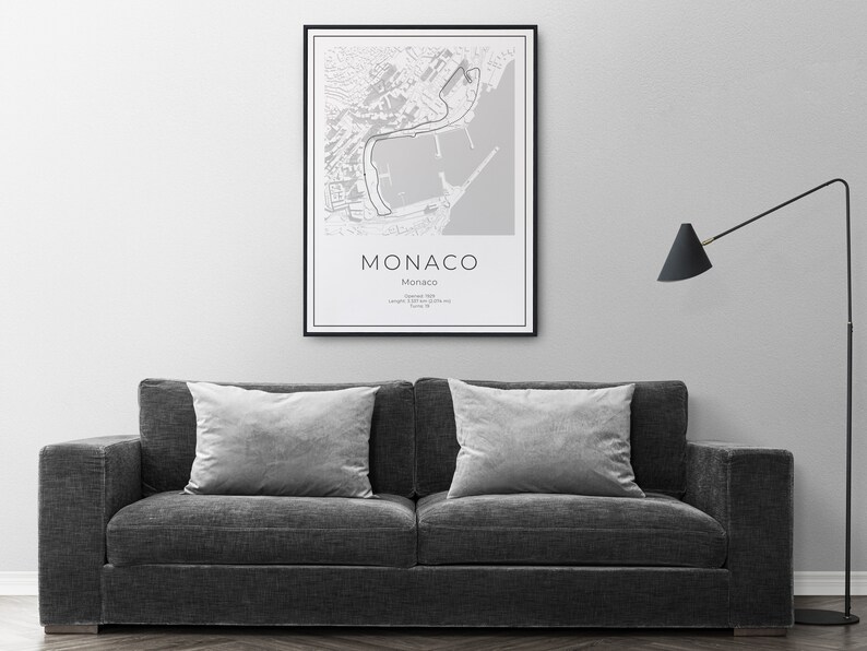 K&ouml;nnte beinhalten: Eine gerahmte Schwarz-Wei&szlig;-Karte von Monaco h&auml;ngt an einer grauen Wand &uuml;ber einem dunkelgrauen Sofa mit zwei grauen Kissen. Die Karte zeigt das Wort "Monaco" in einer klaren Schriftart.