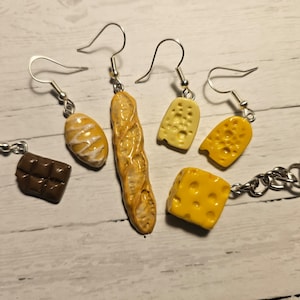 Peut inclure: Cinq paires de boucles d'oreilles avec des breloques miniatures en forme d'aliments, dont une baguette, un petit pain, une barre de chocolat et deux types de fromage différents. Les boucles d'oreilles sont argentées et ont une fermeture à crochet. Les breloques sont en pâte polymère et peintes dans des couleurs réalistes.