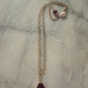 May include: A gold-toned chain necklace featuring a triangular purple gemstone pendant at the base. The necklace includes a lobster claw clasp closure adorned with a small, translucent purple heart-shaped bead and a smooth, pale pink oval bead.