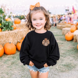 Peut inclure: Un sweat-shirt noir avec un motif monogramme. Le monogramme présente les lettres "AWA" dans un motif à carreaux et léopard, avec un détail de nœud. L'enfant porte un short en jean et un nœud orange.
