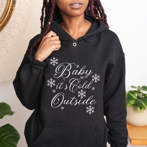 Puede incluir: Sudadera con capucha negra con el texto "Baby it's Cold Outside" en letras blancas cursivas y diseños de copos de nieve.