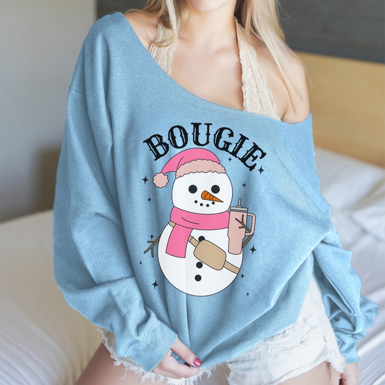 May include: A light blue off-the-shoulder sweatshirt with the text "BOUGIE" in black lettering and a cartoon snowman holding a pink drink. The snowman is wearing a pink scarf and a pink hat.