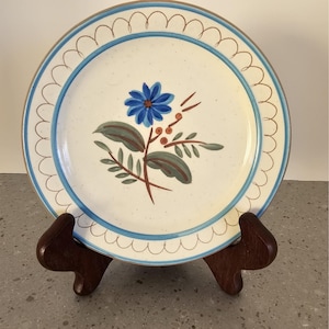 Könnte beinhalten: Ein runder, cremefarbener Keramikteller mit blauem Blumendesign in der Mitte. Der Teller hat einen blauen, gewellten Rand und einen braunen Rand. Er wird auf einem dunklen braunen Holz-Tellerständer präsentiert. Der Teller hat einen Durchmesser von ca. 25 cm.