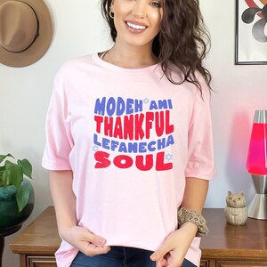 May include: Pink t-shirt with a graphic design that says "Modeh Ani Thankful Lefaneicha Soul" in blue and red letters with a star of David on either side of the text.
