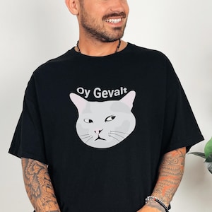 May include: Black t-shirt featuring a grumpy-looking gray cat face with the text "Oy Gevalt" above it. The cat graphic has a minimalist design. The shirt is a casual, oversized style.