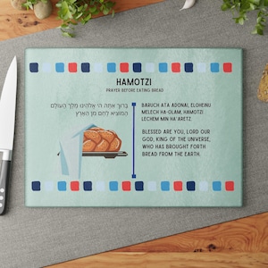 Puede incluir: Una tabla de cortar rectangular de vidrio con el texto "HAMOTZI PRAYER BEFORE EATING BREAD" y una oración en hebreo e inglés. La tabla presenta un gráfico de pan y cuadrados decorativos azules, rojos y blancos.