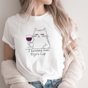 Może przedstawiać: Biała koszulka z kreskówkowym kotem trzymającym kieliszek czerwonego wina. Kot ma zrzędliwą minę. Tekst "I knocked over Elija's Cup" jest wydrukowany pod ilustracją kota. Koszulka jest wykonana z miękkiego materiału.