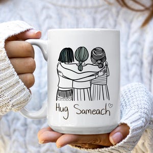 May include: White ceramic mug featuring a black and white illustration of three figures embracing. The mug has the words "Hug Sameach" in a handwritten font, with a small heart symbol. The mug is held by a person.