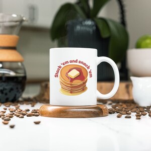 May include: White ceramic coffee mug with a brown wooden coaster. The mug has a graphic of a stack of pancakes with a pat of butter and syrup. The text on the mug reads "Stack 'em and snack 'em".