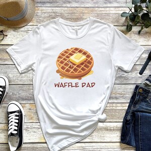 K&ouml;nnte beinhalten: Wei&szlig;es T-Shirt mit einer Cartoon-Illustration einer Waffel mit Butter und Sirup. Der Text "Waffle Dad" ist unter der Waffel aufgedruckt.