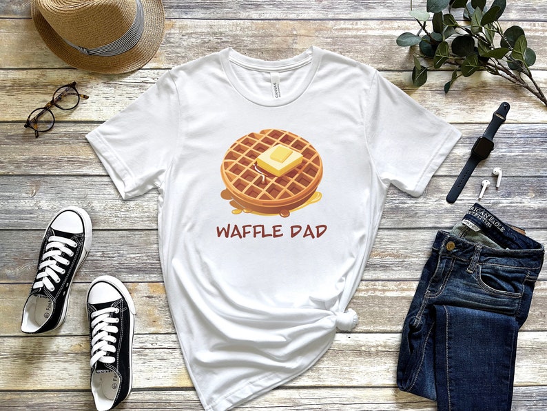 K&ouml;nnte beinhalten: Wei&szlig;es T-Shirt mit einer Cartoon-Illustration einer Waffel mit Butter und Sirup. Der Text "Waffle Dad" ist unter der Waffel aufgedruckt.