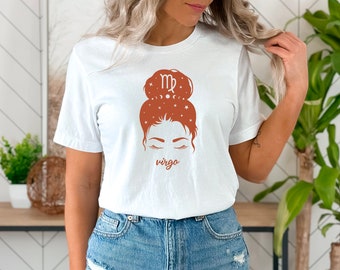 Puede incluir: Camiseta blanca con un gráfico marrón de una mujer con un moño. El moño tiene el símbolo del zodíaco Virgo y estrellas. El texto "virgo" está debajo del gráfico.