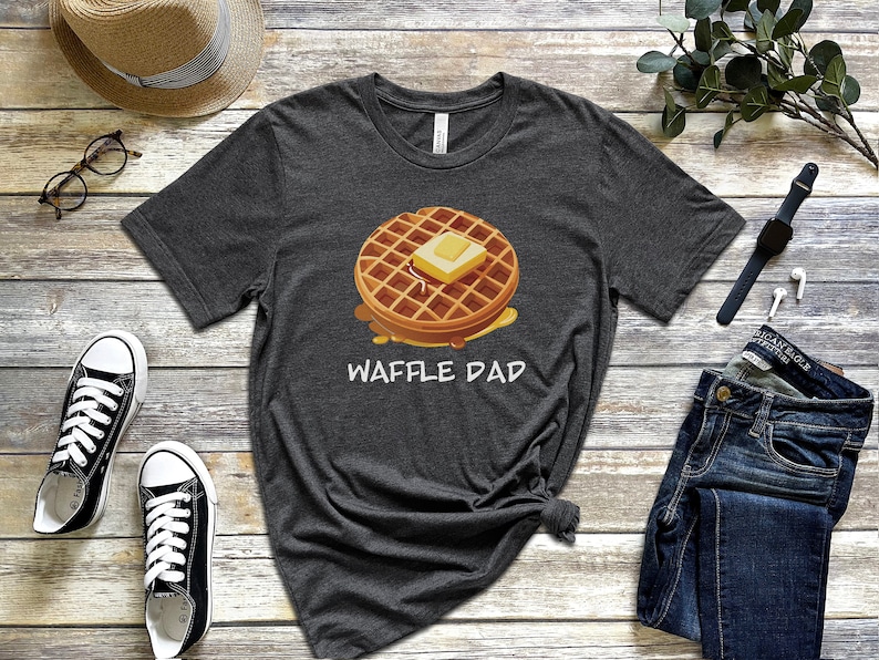 K&ouml;nnte beinhalten: Ein dunkelgraues T-Shirt mit einer Cartoon-Waffel mit Butter darauf und dem Text "Waffle Dad" auf der Vorderseite.
